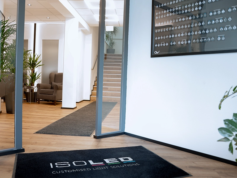 Moderner Eingangsbereich mit Glastüren, Holzfußboden, grauen Teppichen und Sitzbereich mit Sesseln und Pflanzen; Fußmatte mit Aufschrift ISOLED CUSTOMISED LIGHT SOLUTIONS.